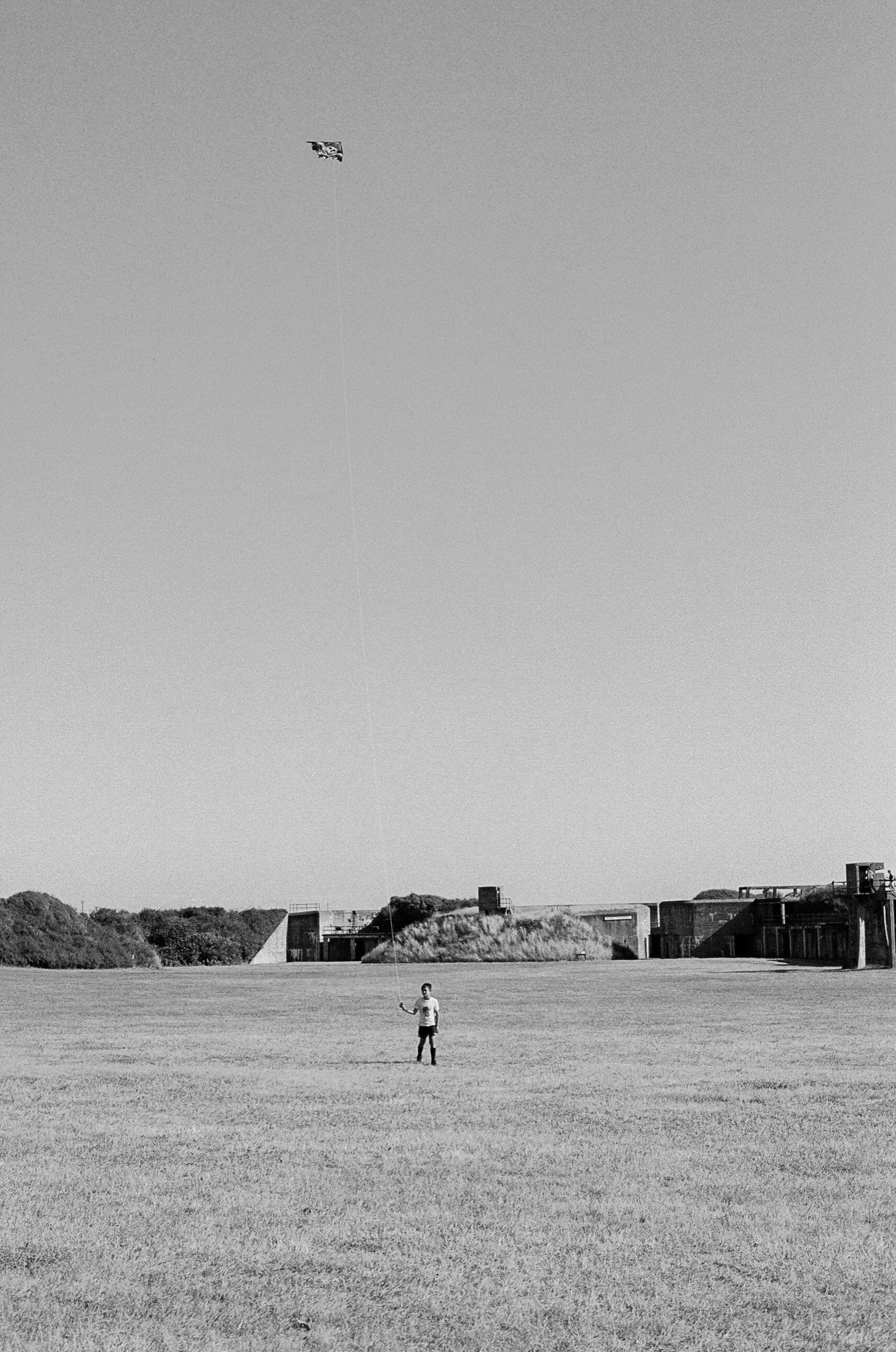 A boy alone in a wide field, flying a small pirate-flag kite high above an old coastal fortification